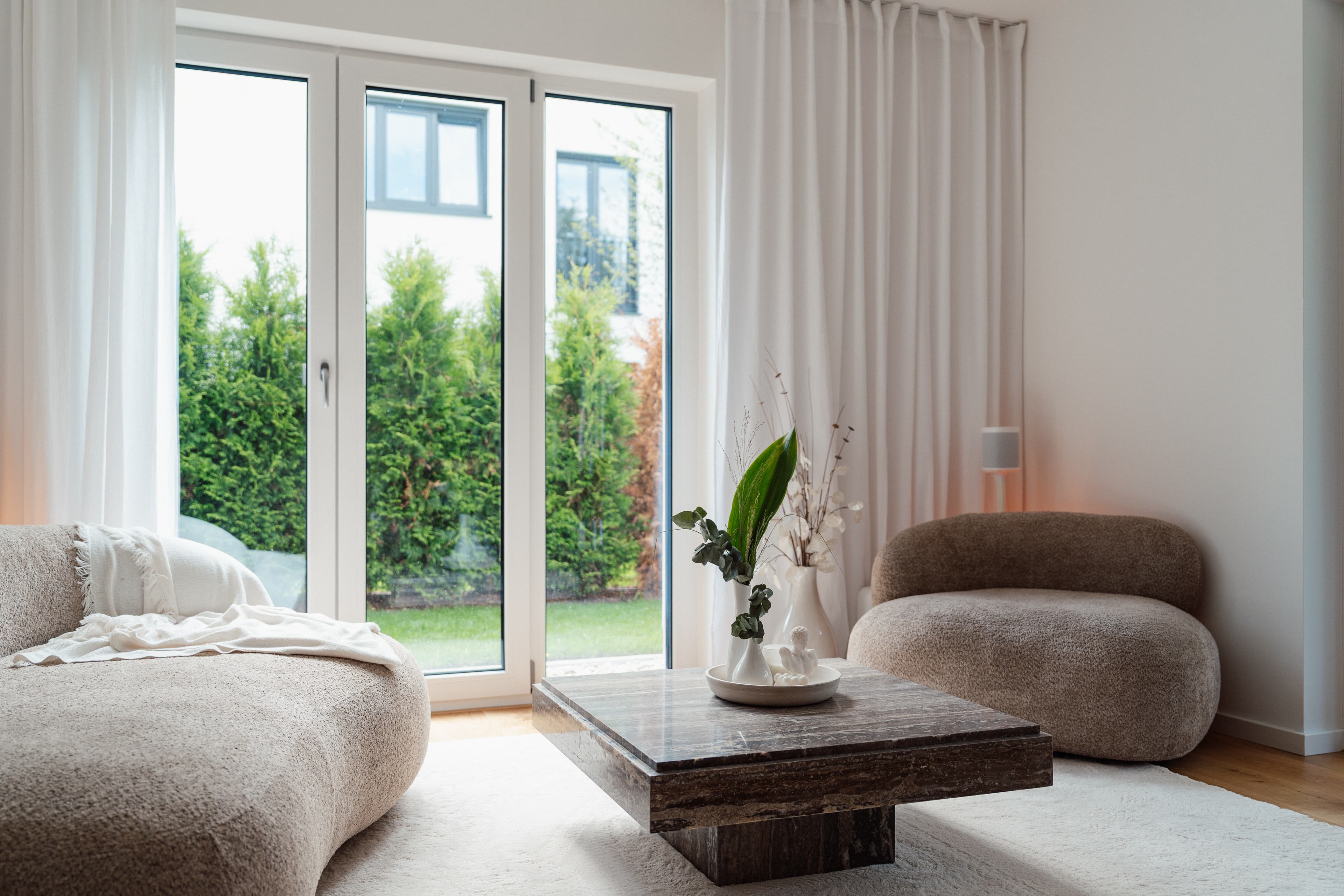 Helles Wohnzimmer mit bodentiefen Terrassentüren, runden Stoffsofas, Vorhängen und dunklem Marmor-Couchtisch