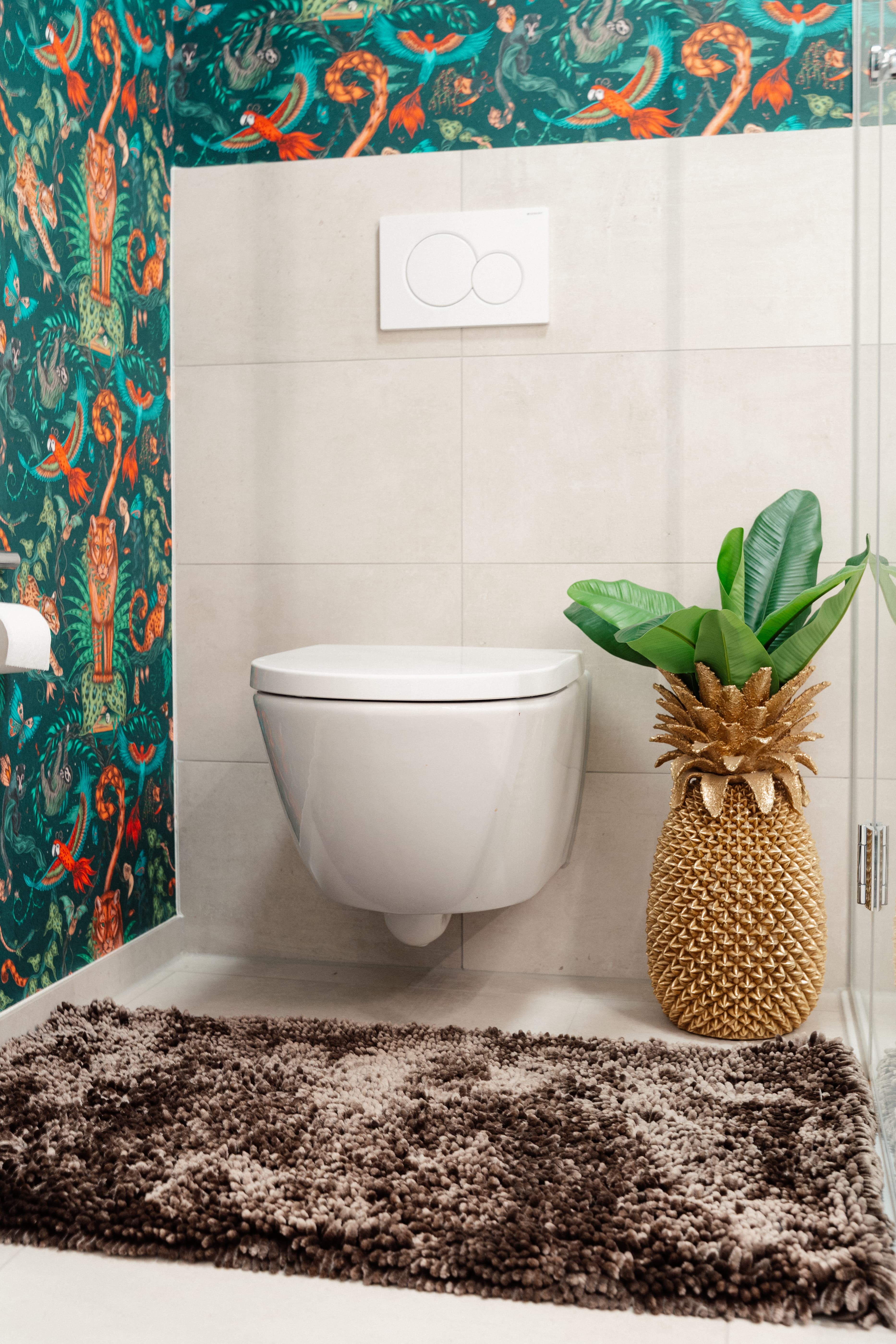 Gäste-WC mit wandhängendem WC, helle Fliesen; tropische Tapete, brauner Teppich und Pflanze im goldenen Ananas-Topf neben Glasduschwand
