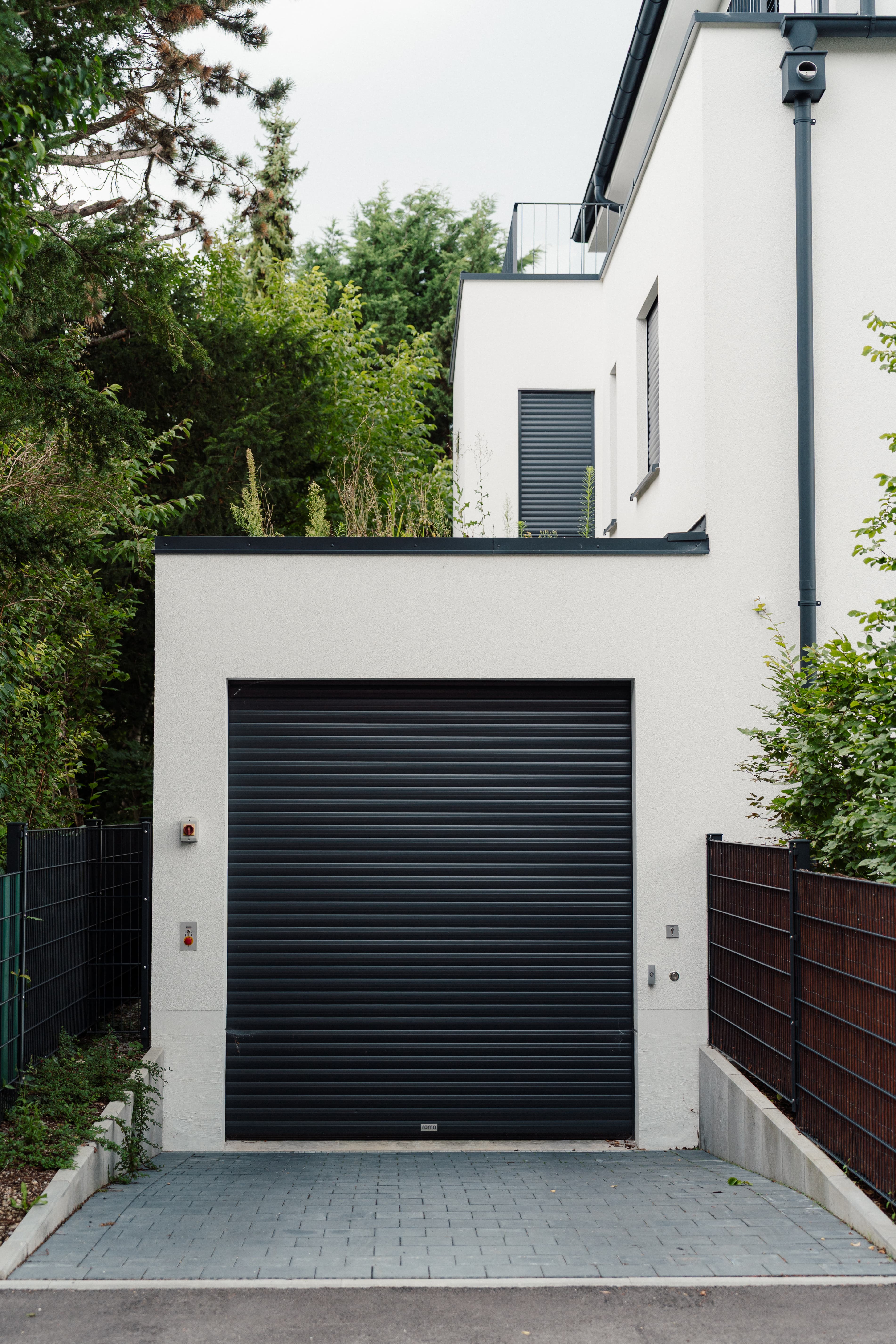 Garage mit schwarzem Rolltor an modernem Wohnhaus; gepflasterte Einfahrt, seitliche Zäune und Begrünung