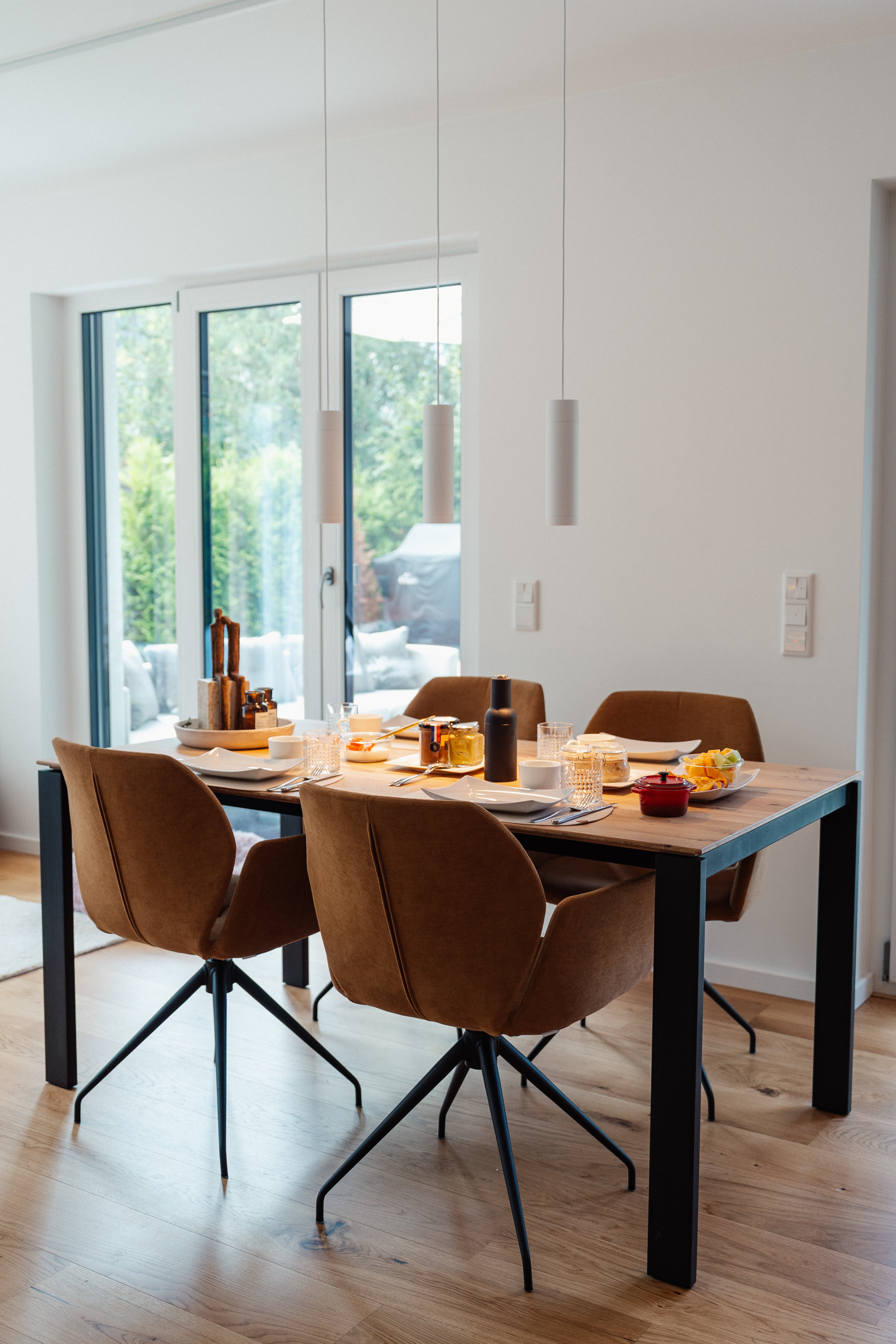 Essbereich mit Holztisch und vier braunen Stühlen; gedeckter Frühstückstisch, bodentiefe Terrassentür und Pendelleuchten