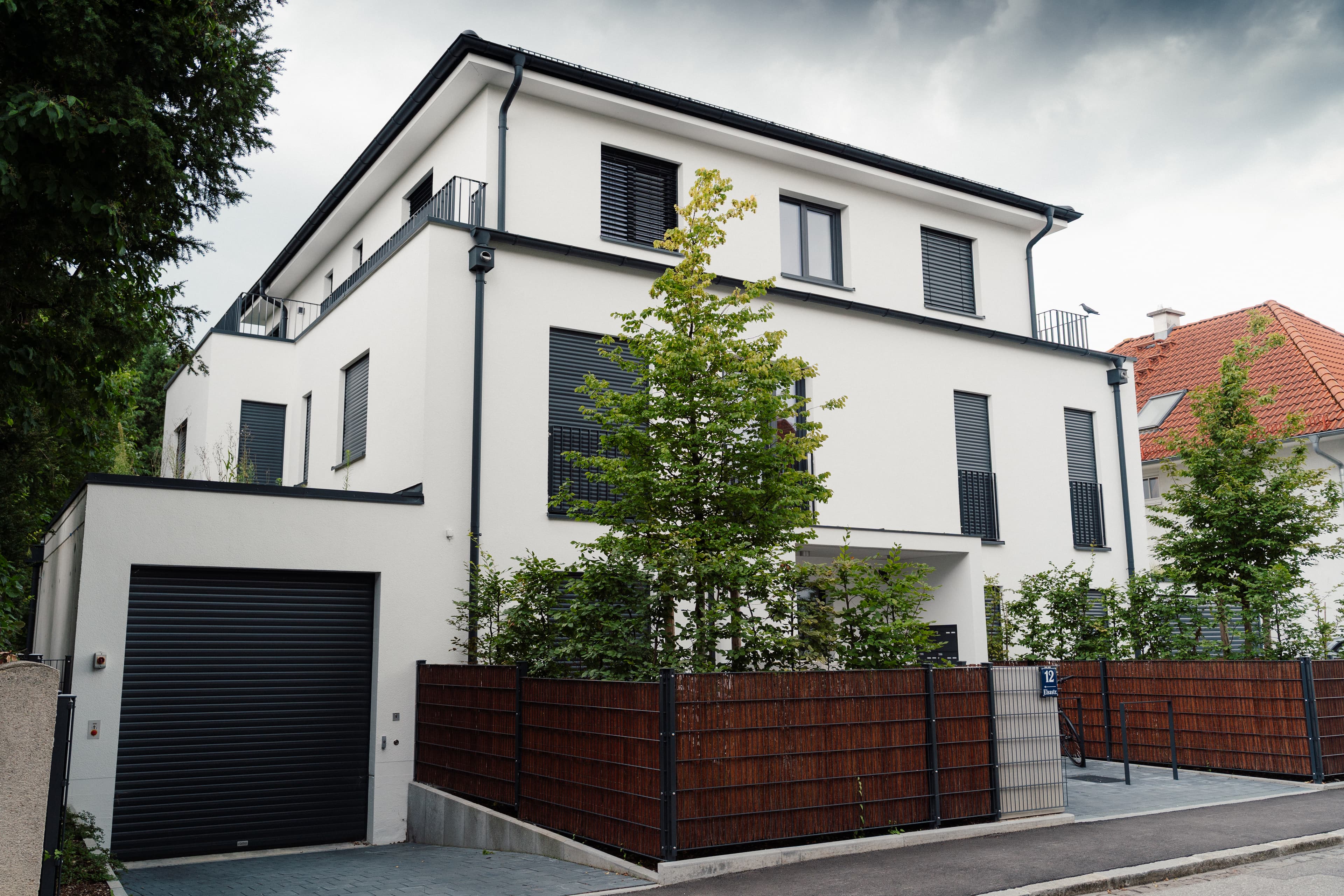 Elsastraße 12 – modernes Mehrfamilienhaus mit weißer Fassade, grauen Rollläden und Garage, Straßenecke mit Zaun und jungen Bäumen